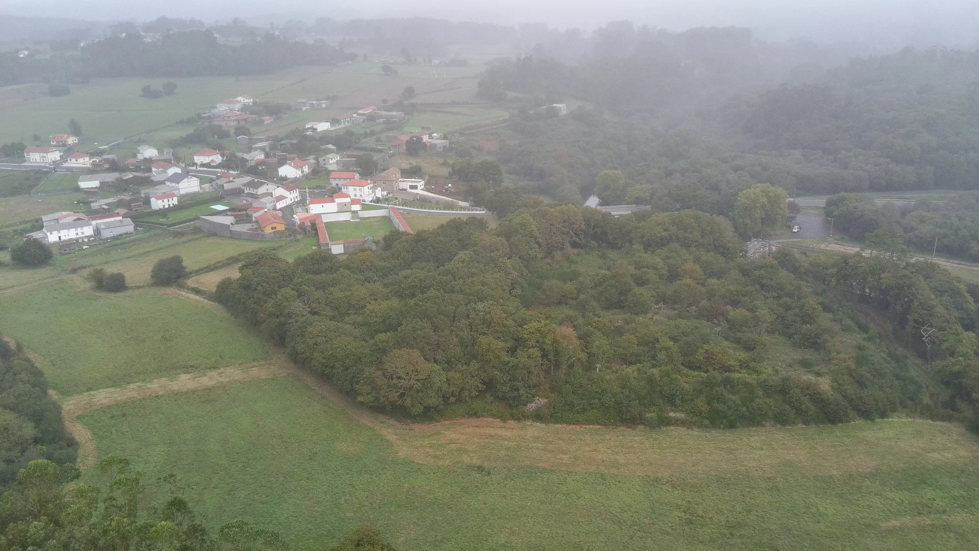 Vista aérea do castro de Brandomil cuberto de arboredo, coa aldea e o cemiterio ao fondo entre néboa